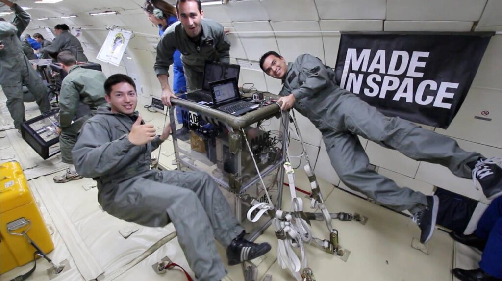 Three men in grey flight suits working with scientific equipment in zero gravity, surrounded by cables and a Made In Space banner.