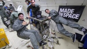 Three men in grey flight suits working with scientific equipment in zero gravity, surrounded by cables and a Made In Space banner.
