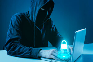 A hooded figure uses a laptop, with a glowing digital padlock