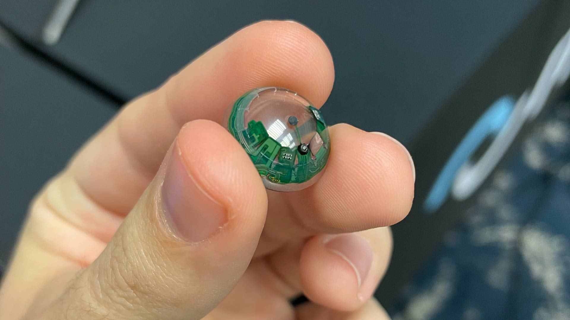 Fingers holding a small, clear plastic sphere containing intricate green circuitry.