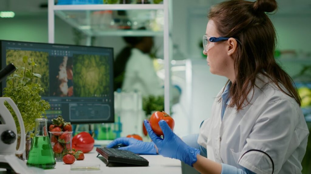 A female scientist examines a tomato in a laboratory, surrounded by plants and research equipment.